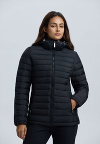 Frau in einer schwarzen gesteppte Jacke mit hohem Kragen und Reißverschlusstaschen, die vor einem schlichten hellen Hintergrund steht.