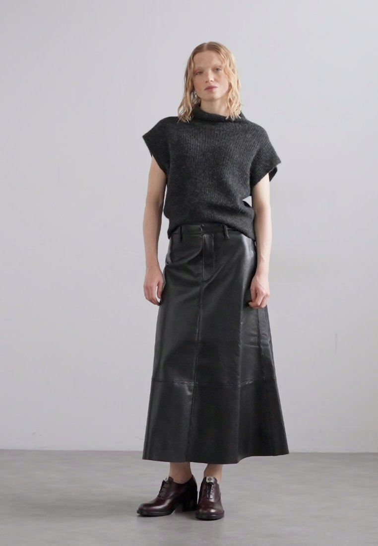 Jupe midi en cuir noir associée à un haut en maille texturée foncé à manches courtes. Le modèle porte des chaussures bordeaux avec un talon épais.