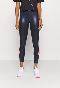 Legginsy w ciemnym granacie z błyszczącym wykończeniem, z różowymi akcentami po bokach. Połączone z białymi sneakersami i czarną bluzką.