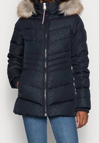Granatowa kurtka puchowa z kapturem wyściełanym futrem, w geometryczny wzór, z zamkiem z przodu, ściągaczami przy mankietach oraz bocznymi kieszeniami. Izolowana dla zapewnienia ciepła.