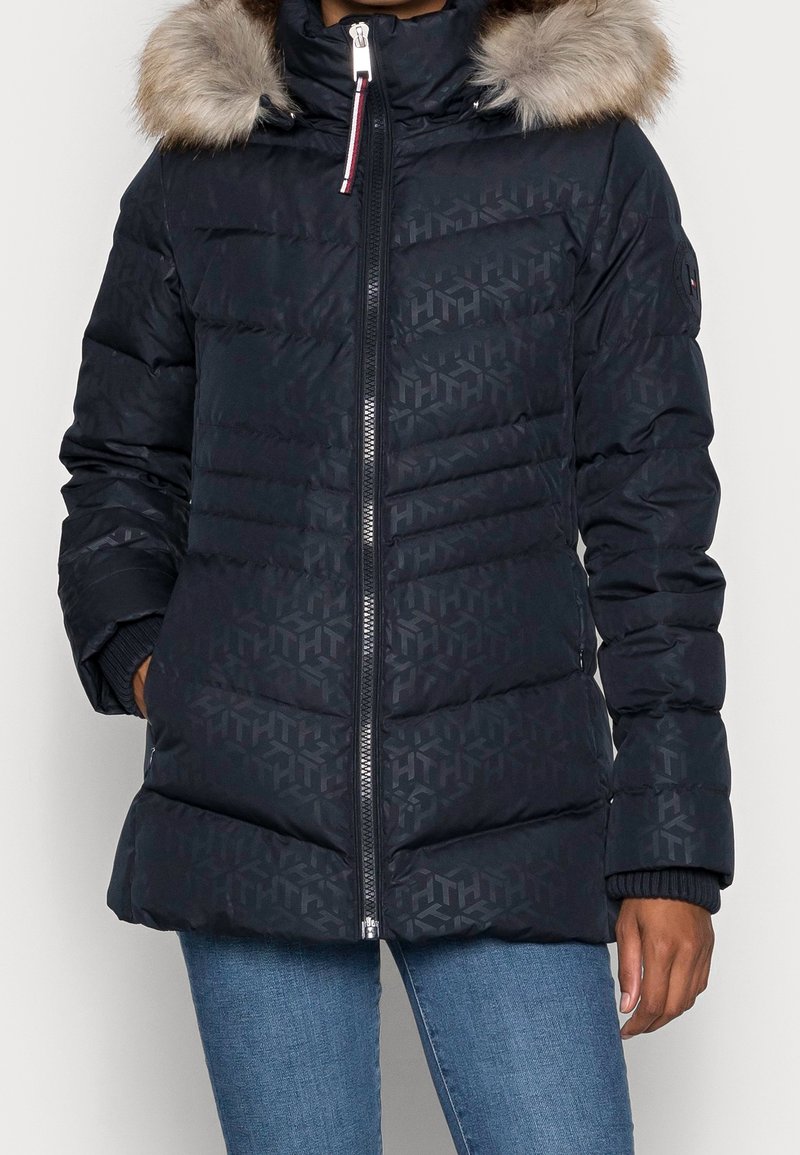 Granatowa kurtka puchowa z kapturem wyściełanym futrem, w geometryczny wzór, z zamkiem z przodu, ściągaczami przy mankietach oraz bocznymi kieszeniami. Izolowana dla zapewnienia ciepła.