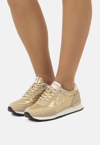 Baskets d'athlétisme dorées avec un mélange de textures brillantes et en cuir suédé, des lacets fins, une semelle rembourrée et un logo blanc sur la languette.