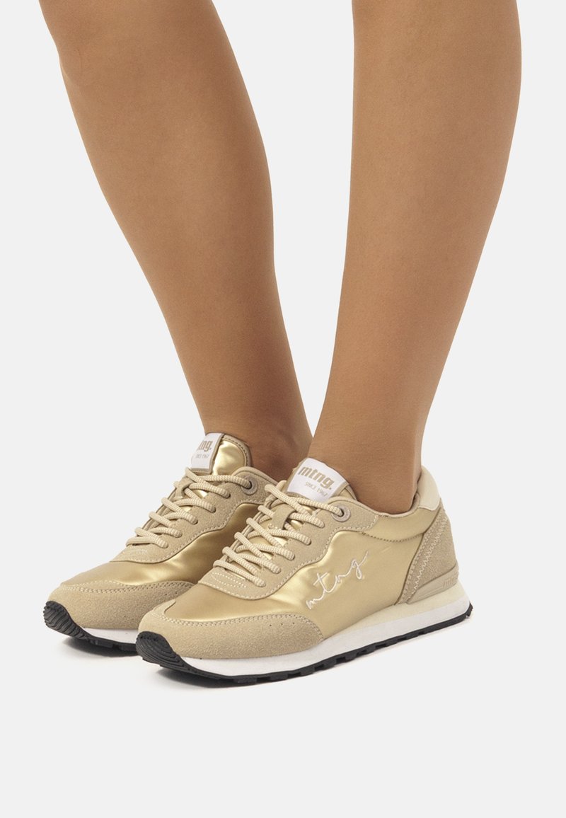 Baskets d'athlétisme dorées avec un mélange de textures brillantes et en cuir suédé, des lacets fins, une semelle rembourrée et un logo blanc sur la languette.