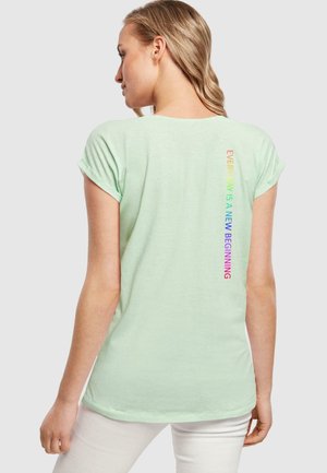 Mintgrøn t-shirt med korte opbollerede ærmer, der har et lodret flerfarvet tekstdesign på ryggen, som lyder: "HVER DAG ER EN NY BEGINDELSE."