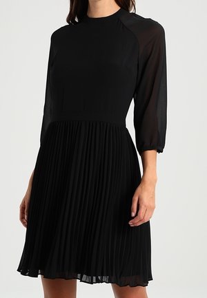 Femme portant une robe noire longueur genou avec des manches trois-quarts transparentes et une jupe plissée.