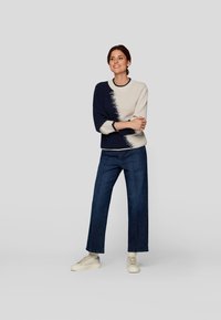 Strickpullover mit einem Verlaufdesign in Marineblau und Creme, mit Dreiviertelärmeln. Kombiniert mit weit geschnittenen, dunkelblauen Jeans und hellen Sneakers.