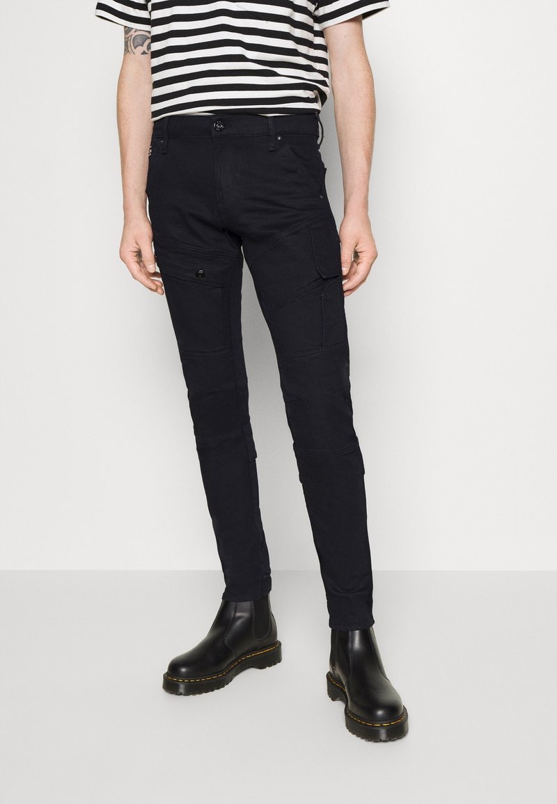 Pantalones cargo negros con múltiples bolsillos, ajuste slim, tejido ligeramente texturizado. Combinados con botas negras hasta el tobillo y una camisa a rayas.
