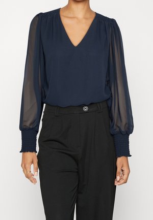 Femme portant un chemisier bleu marine à col en V avec des manches longues transparentes et des poignets smockés, associé à un pantalon noir taille haute.