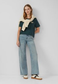 T-shirt vert foncé avec des détails brodés, jeans mom bleu clair, pull en tricot crème drapé sur les épaules et baskets élégantes.