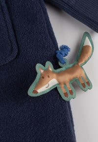 Marineblaue Fleecjacke mit strukturierten Oberflächen und einem braunen, illustrierten Fuchs-Patch, verziert mit grünen Akzenten und einer blauen Stoffschlaufe.