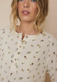 Hellcremefarbenes Hemd zum Knöpfen aus strukturiertem Stoff mit kleinen Blumenmustern in Pink, Gelb und Blau, mit metallischen Knöpfen versehen.