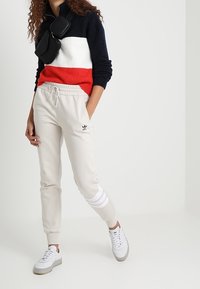 Mörkblå, röd och vit tröja med halvdragkedja; krämfärgade sweatpants med tre vita ränder; svart crossbody-väska; vita sneakers med grå detaljer.