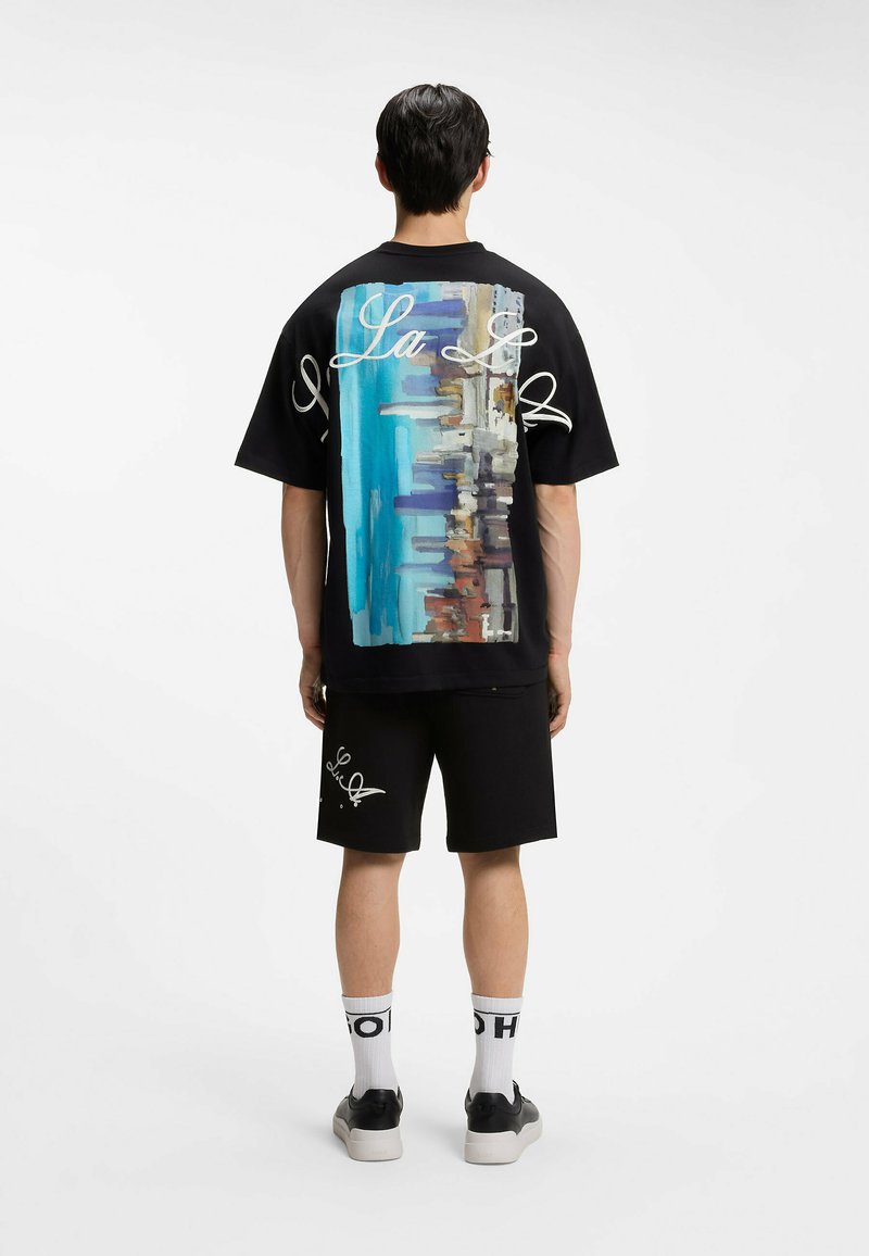 Man wearing black shorts and a black t-shirt with a colorful cityscape and white script text on the back, standing facing away.
