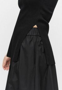 Czarny sweter z prążkowanej dzianiny z bocznymi rozcięciami, zestawiony z czarną plisowaną spódnicą. Tkaniny mają gładką fakturę i prosty design.