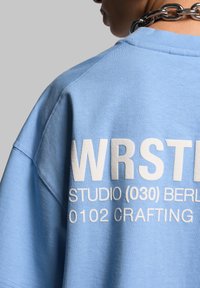 Vaaleansininen oversized-sweatshirt, joka on valmistettu puuvillasta. Takana valkoisella painettuna teksti: "WRSTL STUDIO (030) BERLIN 0102 CRAFTING".