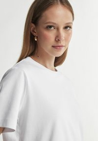 Hvid bomulds t-shirt med rund hals, korte ærmer og en afslappet pasform, kombineret med enkle guldfarvede stud øreringe. Glat tekstur.