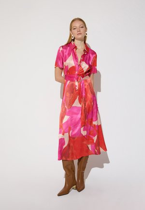 Femme aux longs cheveux blonds portant une robe midi ceinturée à motifs rose, rouge et beige, des bottes marron montant jusqu'aux genoux et des boucles d'oreilles blanches, debout devant un fond blanc.
