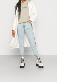 Manteau blanc long sur un sweat à capuche gris, jeans bleu clair et bottes de combat noires à lacets. Présente un design simple avec une coupe décontractée et droite.