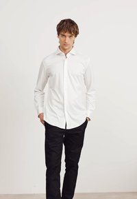 Camicia bianca a maniche lunghe con colletto, abbinata a pantaloni neri, in piedi davanti a uno sfondo chiaro. Texture liscia, vestibilità su misura.