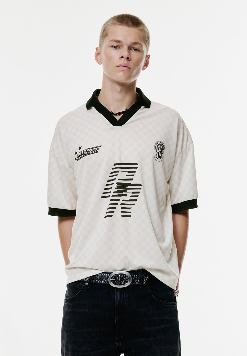 Jonge man in een wit geruit sportshirt met zwarte kraag en logo's, zwarte riem met zilveren gesp en donkere broek, staand met zijn handen achter zijn rug.