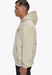Beige hoodie with a front pocket, a drawstring hood, and ribbed cuffs. Features a logo embroidered in brown on the chest.
