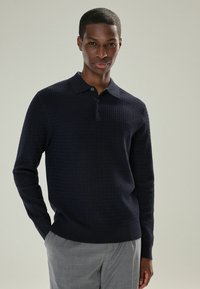 Marineblaues, langärmliges Poloshirt mit strukturiertem Muster, 3-Knopf-Leiste und gerippten Bündchen, kombiniert mit hellgrauen Hosen.