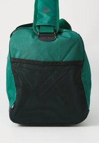 Grønn stoff skulderbag med et teksturert svart nettpanel, justerbar stropp og grå logo aksent på toppen. Har glidelås-lukking.
