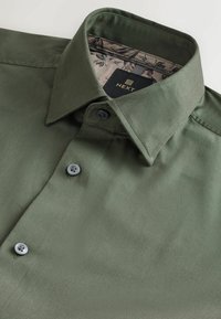 Olivgrünes Button-up Hemd mit Kragen, ausgestattet mit zwei grauen Knöpfen, aus glattem Stoff und mit einem floralen Muster auf der Innenseite des Kragens. Markenetikett sichtbar.