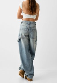 Jeans cargo de mezclilla azul claro, con un corte holgado, múltiples bolsillos traseros y una textura sutilmente desgastada. Combinados con zapatos marrones de suela gruesa.