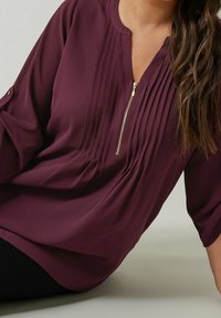 Blouse prune avec des détails plissés, un col zippé et des poignets de manches roulés. Tissu doux et coupe décontractée. Texture lisse.