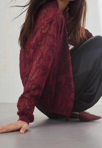 Blouse bordeaux transparente avec motif floral texturé, manches longues avec poignets élastiques, associée à un pantalon noir et des chaussures à bout pointu.