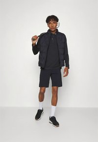 Juoda siuvinėta liemenė dėvima ant juodo polo marškinėlio, dera su tamsiomis šortais. Modelis dėvi juodus sportbačius ir baltas kojines, laiko golfo lazdą.