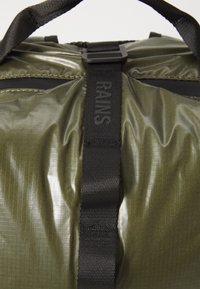 Olive zöld szintetikus táska fényes felülettel, fekete pánttal, amelyen "RAINS" logó található, és textured felülettel, rácsos mintával.