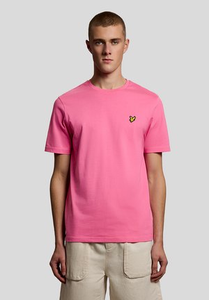 Jaunas vyras su trumpais plaukais, dėvintis paprastą rožinę marškinėlį su mažu geltonu paukščio logotipu ir smėlio spalvos kargo šortus, stovintis priešais pilką foną.