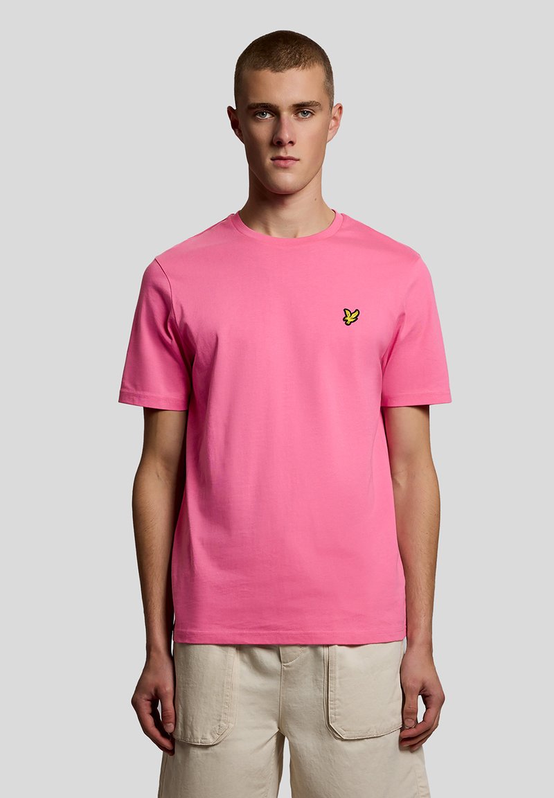 Νεαρός άνδρας με κοντά μαλλιά, φορώντας μια απλή ροζ μπλούζα με λογότυπο μικρού κίτρινου πουλιού και μπεζ cargo σορτς, στέκεται μπροστά σε γκρι φόντο.