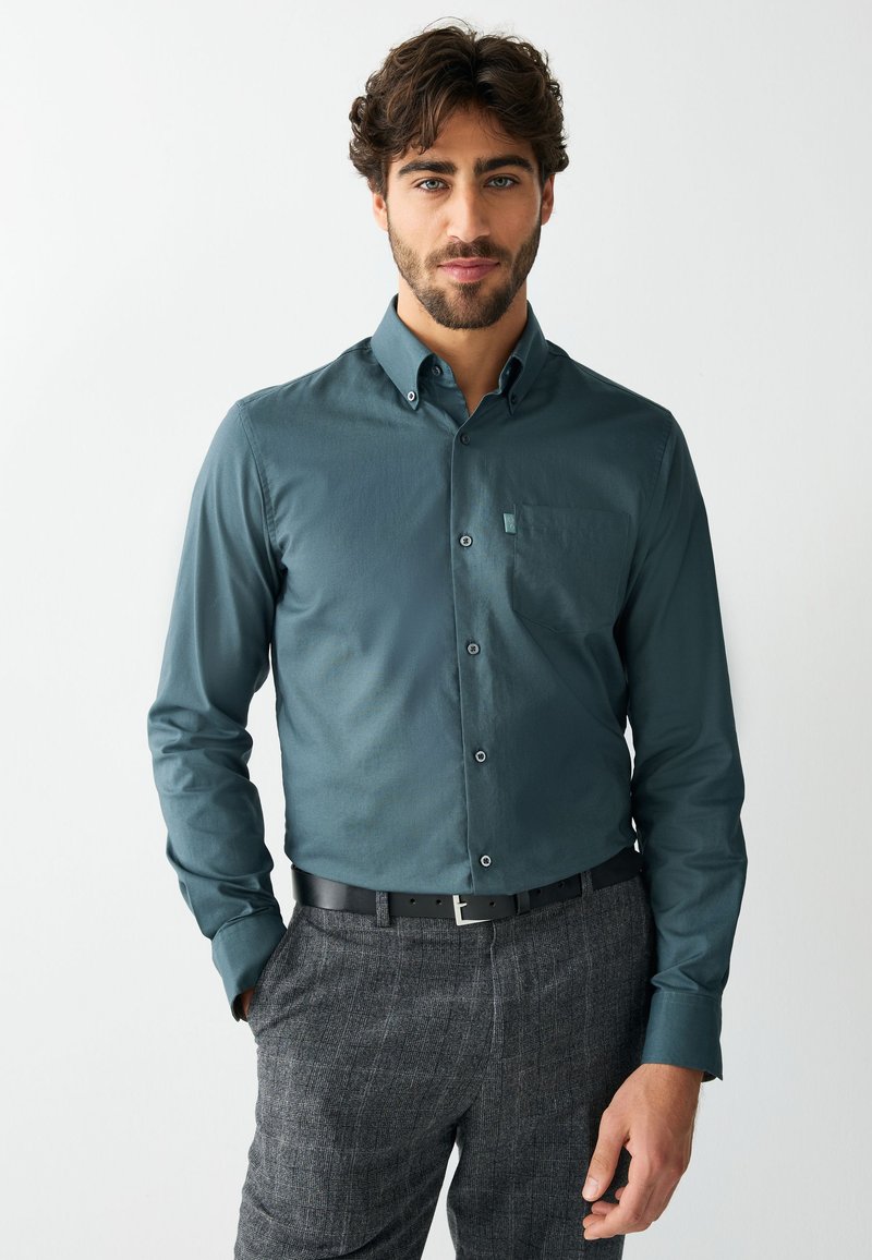 Homme aux cheveux bouclés foncés portant une chemise bleu sarcelle boutonnée et un pantalon gris à carreaux, debout avec une main dans la poche sur un fond uni.