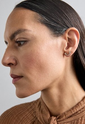 Zelta auskari ar izliekta dizainu, kas ir rotāti ar divām sīkām dārgakmeņiem un rindu mirdzošu akcentu. Modelis ar garām matiem valkā teksturētu brūnu blūzi.