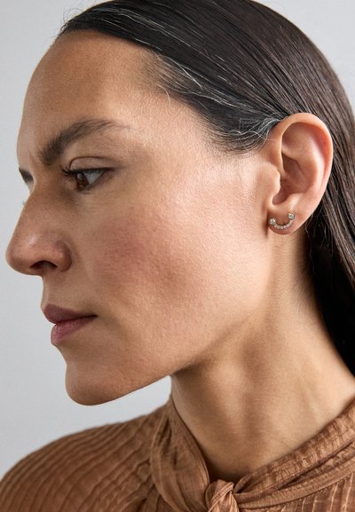 Boucles d'oreilles en or au design courbé, ornées de deux petites pierres précieuses et d'une rangée d'accentuations scintillantes. Un modèle aux longs cheveux porte un haut en brun texturé.