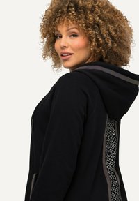 Sudadera con capucha negra que presenta un panel trasero texturizado con un patrón geométrico, cierre frontal con cremallera y acentos en contraste.