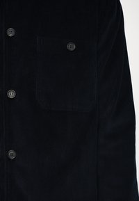 Tummansininen corduroypaita, jossa on napit edessä, kaksi nappia suljettavaksi ja vasemmassa rinnassa tasku, joka on ribbikuvioitu.