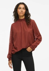 Kastanjebrun blus med långa ärmar, hög volangkrage, zigzag-mönster och smockade ärmslut. Kombinationen med svarta tajta jeans.