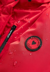 Rød vandtæt jakke med en glat overflade, der har et rundt sort badge med teksten "Eventyret Venter" og vanddråber på overfladen.