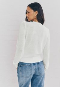Pull en tricot blanc à manches longues et ourlet côtelé, porté avec un jean en denim bleu taille haute. Texture légère avec une coupe décontractée.
