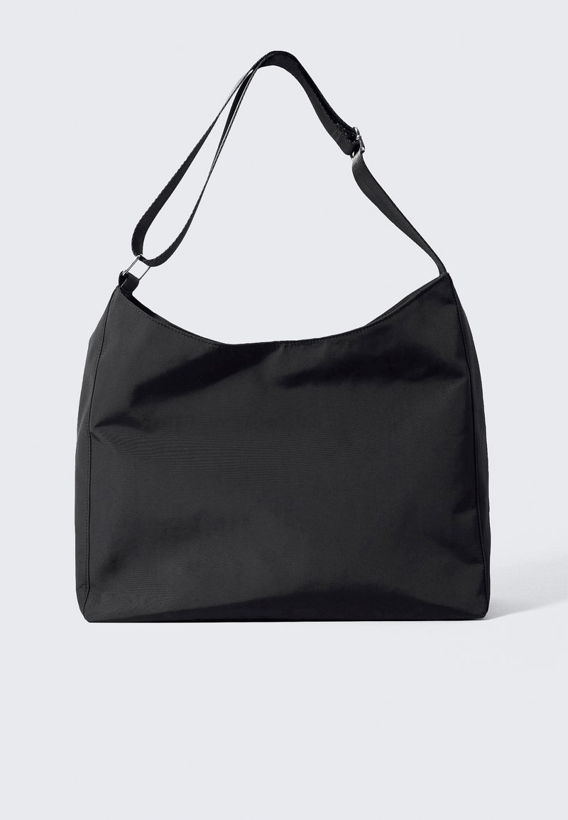 Bolso de hombro negro con correa ajustable, forma rectangular y tela lisa, mostrado sobre un fondo gris claro liso.