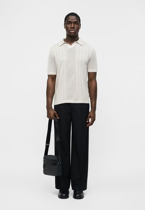 Man staat recht vooruit kijkend, draagt een crèmekleurige korte mouw gebreide polo, zwarte broek met wijde pijpen, zwarte schoenen en houdt een zwarte schoudertas vast.