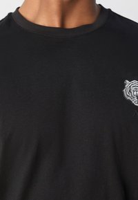 Black cotton t-shirt with a round neckline, featuring a white tiger graphic on the left chest and a subtle brand accent on the hem.