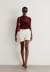 Red and black long-sleeve top, white shorts, black sandals, carrying a red handbag, wearing dark sunglasses, walking against a neutral background.