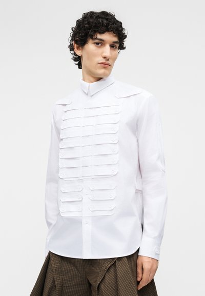 Młody mężczyzna z kręconymi włosami, ubrany w białą koszulę na guziki z warstwowymi paskami tkaniny oraz szerokie spodnie w brązową kratę.