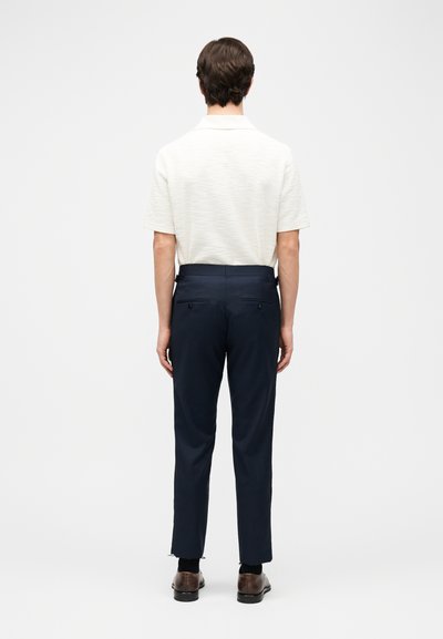 Bărbat care stă cu fața spre spate, purtând un tricou polo alb cu mâneci scurte, pantaloni bleumarin croiți, șosete negre și pantofi maro eleganți.