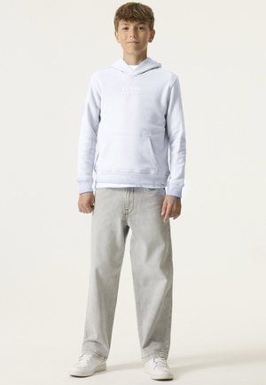 Tienerjongen staat, draagt een witte hoodie met voorzak, lichtgrijze wijde jeans en witte sneakers op een effen achtergrond.